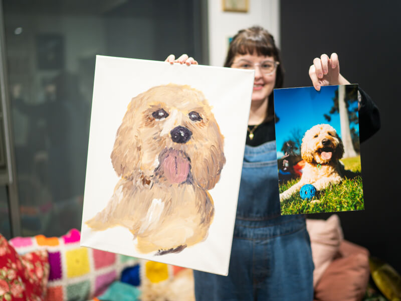 Woman holding up a dog portrait and picture in a painting class